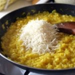 Skillet on the stove filled with risotto alla Milanese, a pile of cheese, and a wooden spoon.
