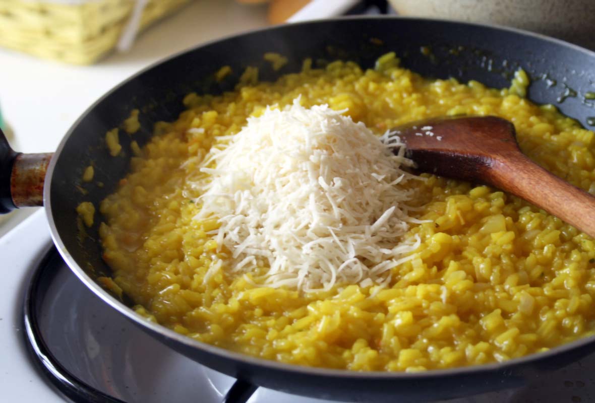 Skillet on the stove filled with risotto alla Milanese, a pile of cheese, and a wooden spoon.