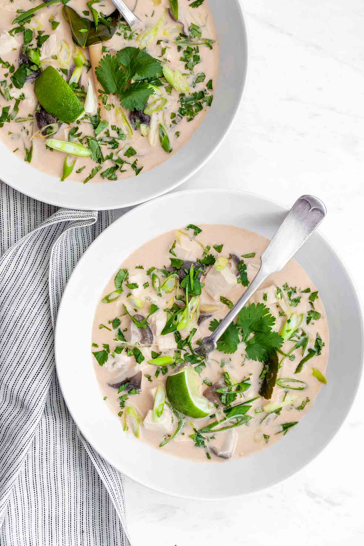 Two bowls of Thai-inspired chicken soup with coconut milk and lemongrass on a striped napkin.