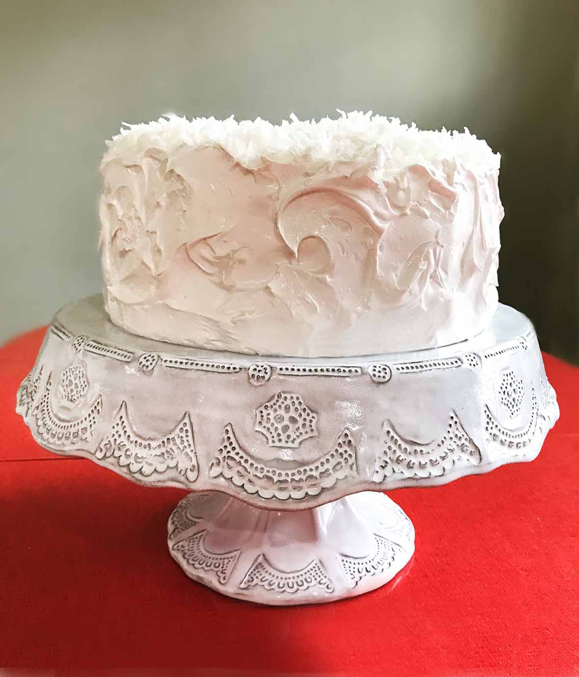 Coconut layer cake with white meringue frosting topped with shredded coconut on a white cake stand on a red table
