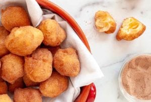 A red pot filled with sonhos dusted with cinnamon sugar and one cut Portuguese doughnut resting beside the bowl