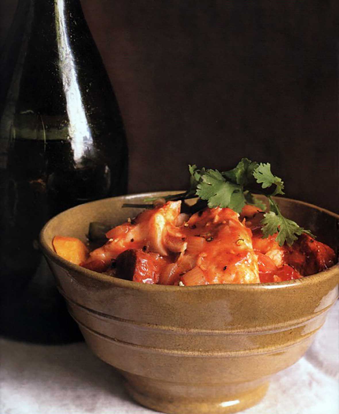 A brown ceramic bowl filled with Portuguese fish chowder, made with fish, potatoes, chouriço, tomatoes, and cilantro.