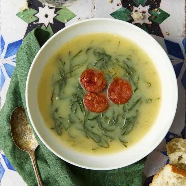 A white bowl of caldo verde, or Portuguese kale soup (green broth) with thinly sliced kale and three chourico coins floating in the center.