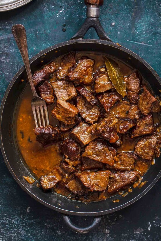 A cast iron skillet with cacoila--or chucks of Portuguese stewed beef, bay leaf, and a fork