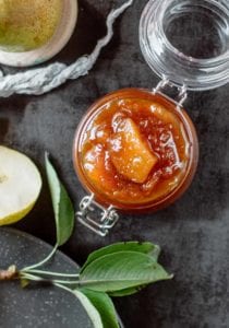 An open jar of pear chutney with a cut pear and some leaves lying beside it.