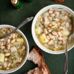 Two white bowls of Portuguese fennel soup with spoons resting inside and crusty bread on the side.
