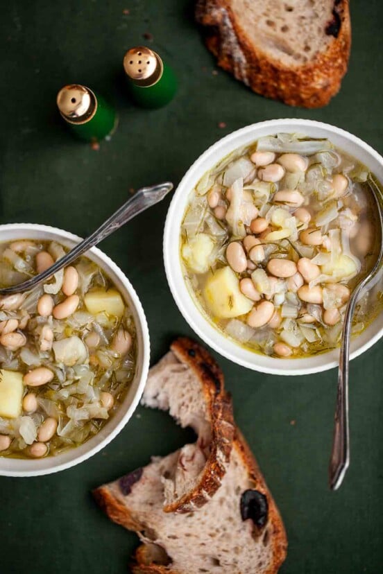 Two white bowls of Portuguese fennel soup with spoons resting inside and crusty bread on the side.