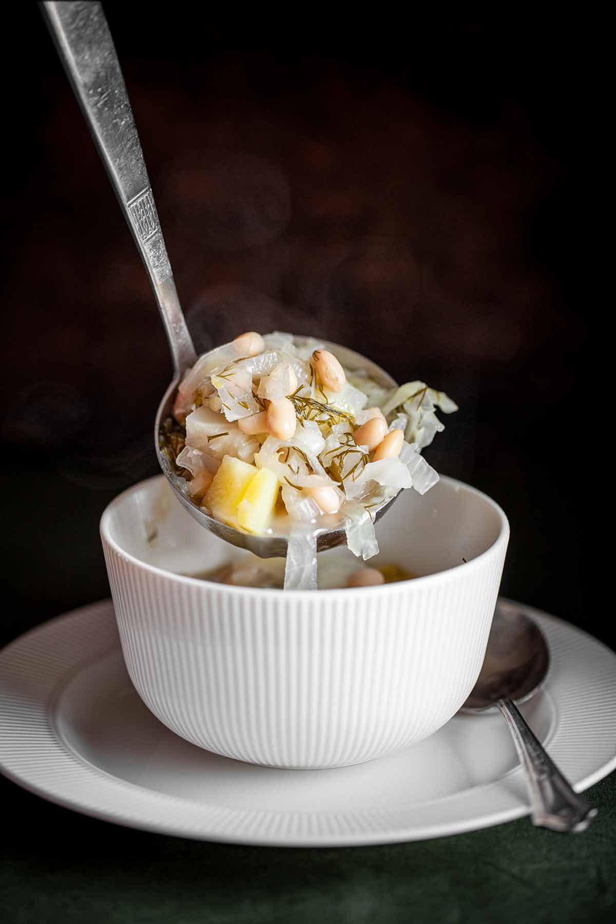 A ladle scooping Portuguese fennel soup into a white bowl on a white plate with a silver spoon beside it.