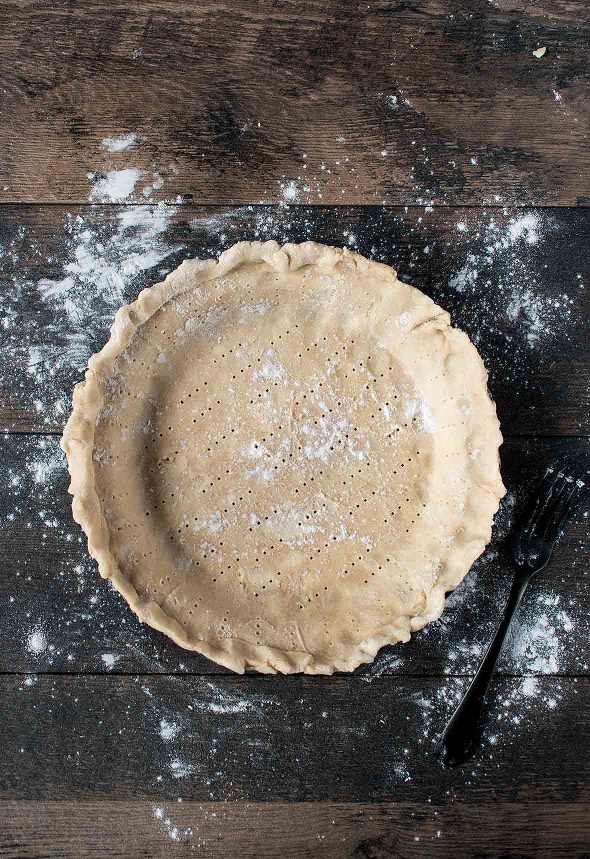 An unbaked lard and butter pie crust, with fork pricks all over on a wooden table.