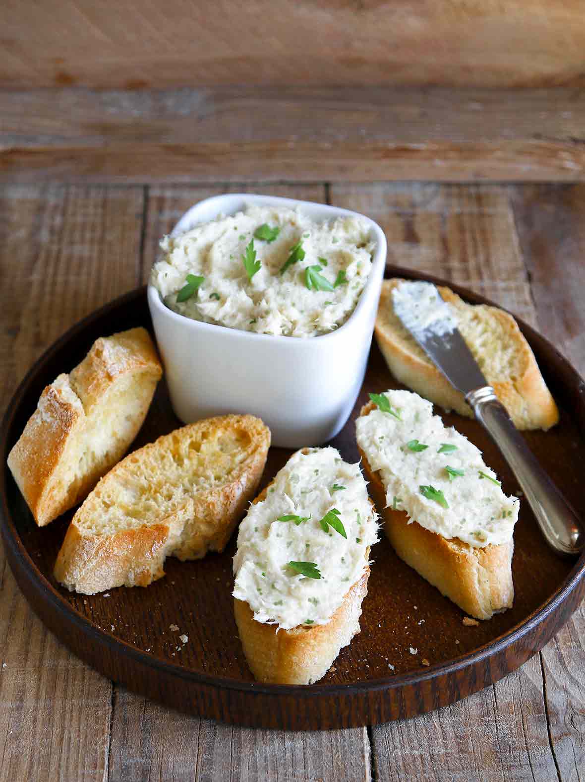 A plate with five baguette slices, a knife, and white bowl filled with fresh cod brandade, garnished with parsley.