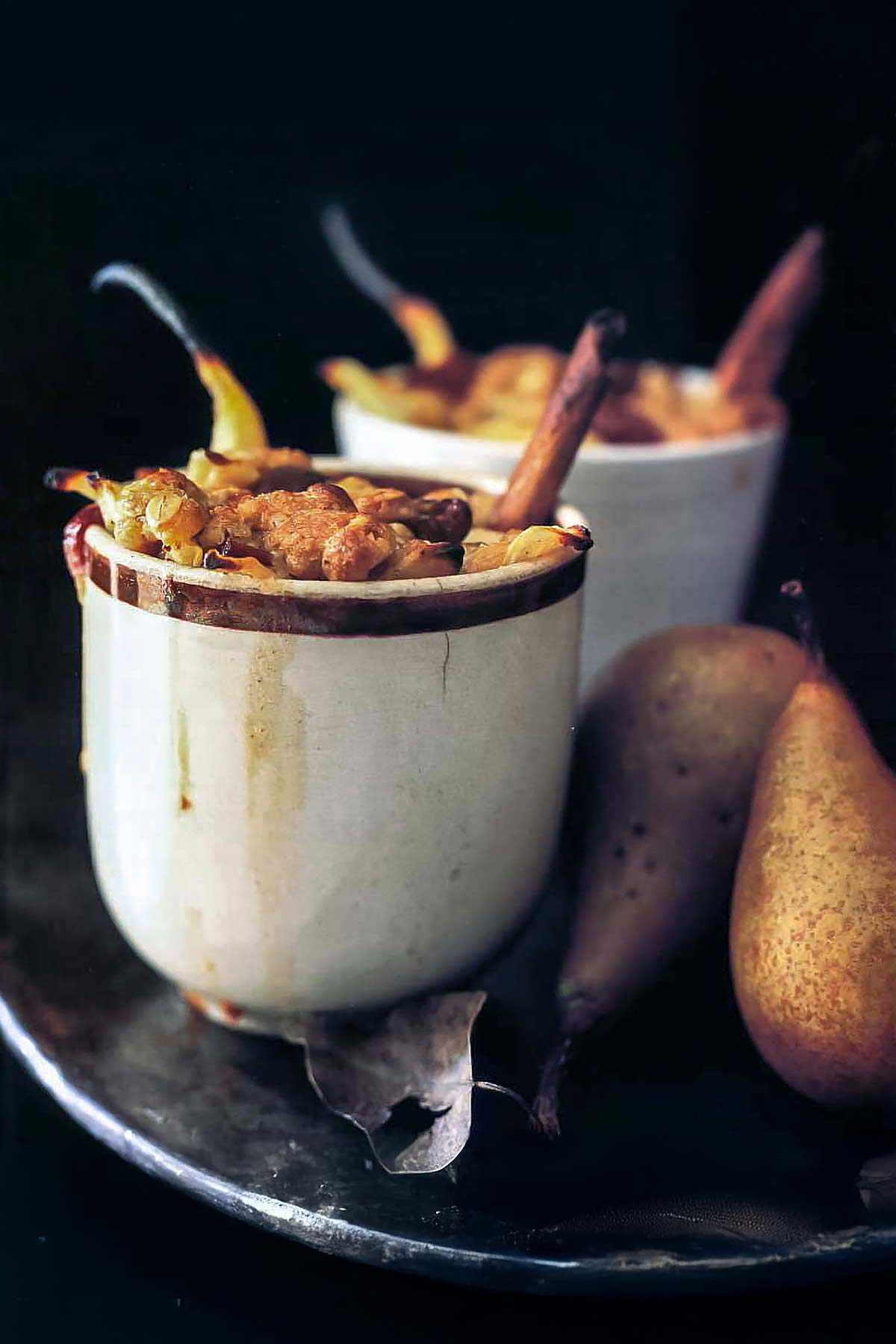 Pear cranberry crumbles in white ramekins with cinnamon sticks and pear stems sticking out, flanked by fresh pears.