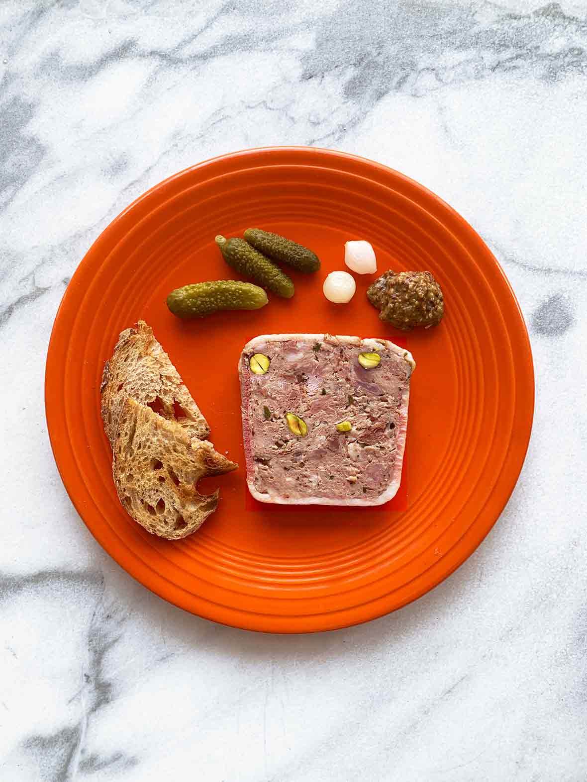 A red plate with a slice of pate de campagne, crostini, cornichons, pickled onions, and a dollop of mustard