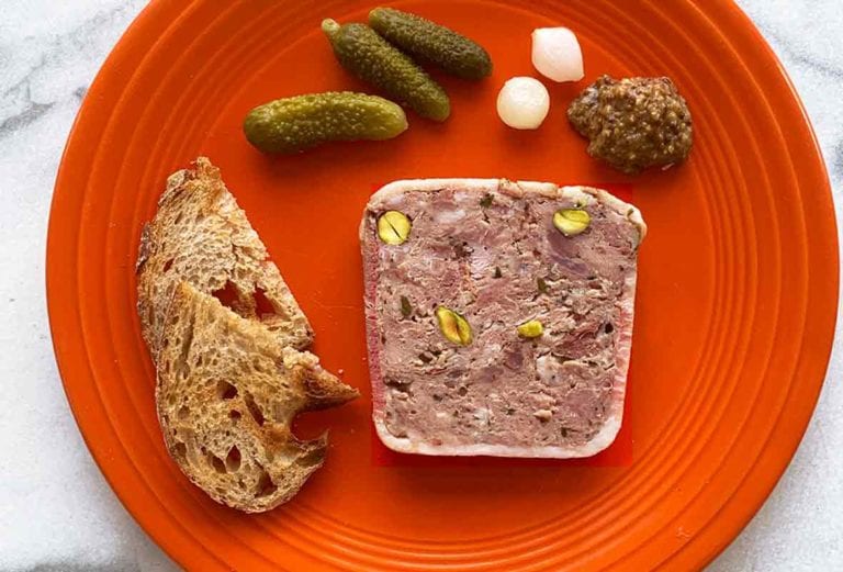 A red plate with a slice of country pate, crostini, cornichons, pickled onions, and a dollop of mustard
