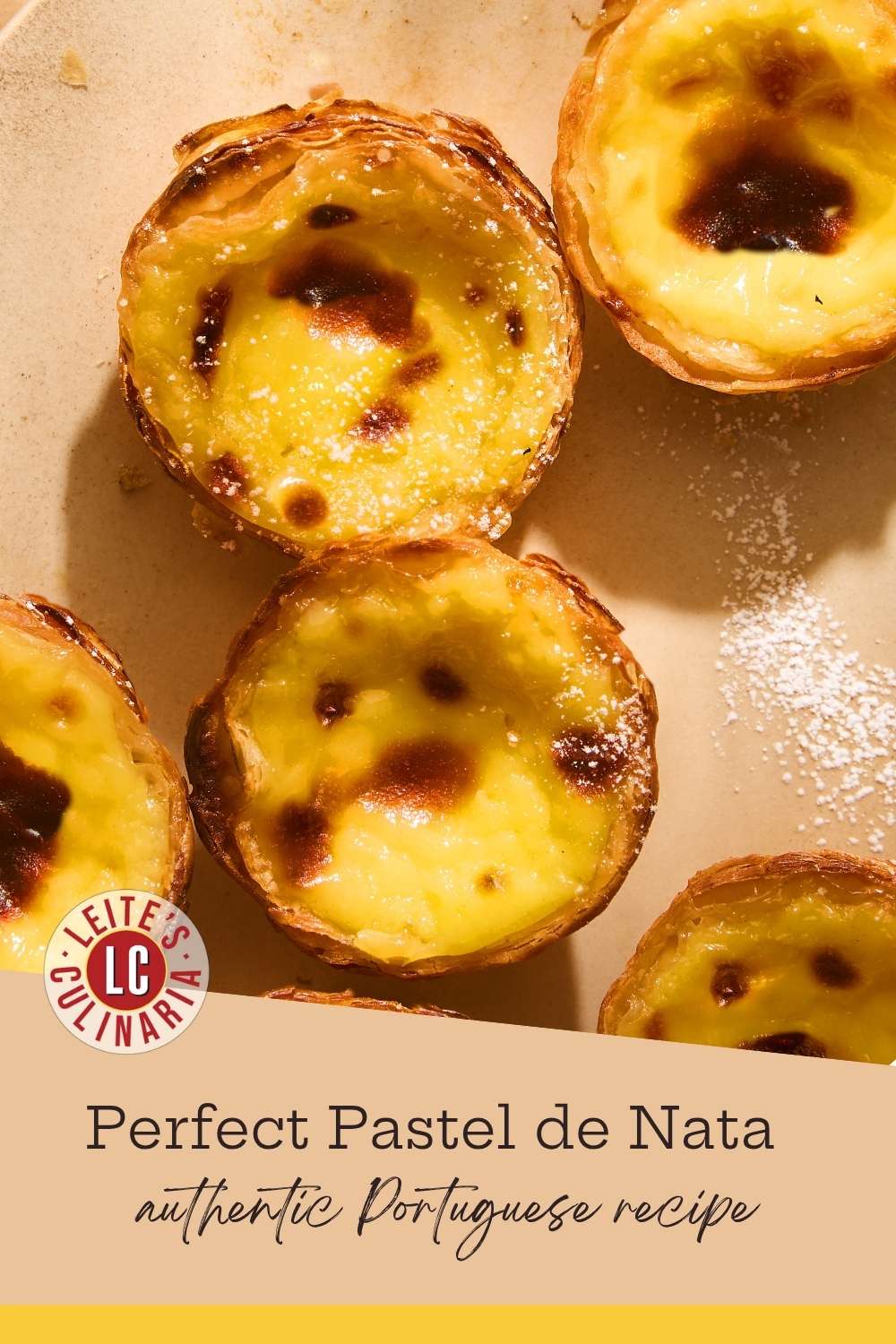Overhead view of several golden-brown Portuguese egg tarts on a cream-colored plate, dusted with powdered sugar.