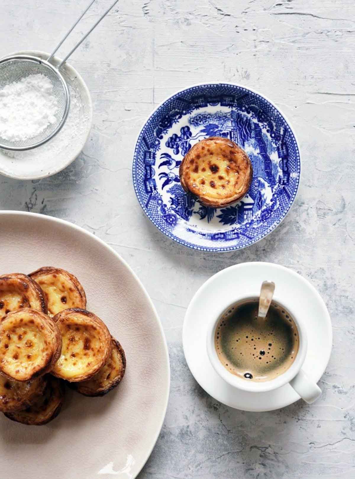 A platter of pasteis de nata near a cup of coffee and a single pastel,
