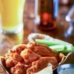 A paper basket filled with classic buffalo wings and celery sticks with a metal cup of dipping sauce beside it.
