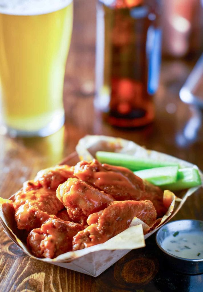 A paper basket filled with classic buffalo wings and celery sticks with a metal cup of dipping sauce beside it.