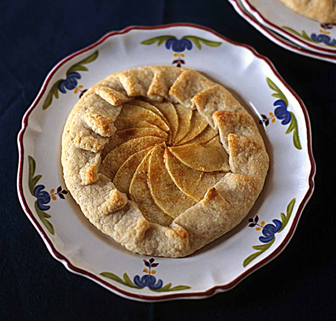 Individual rustic apple tart with the crust folded over a filling of sliced apples on a flowered plate