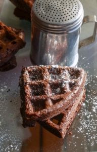 Two dark chocolate waffles on a mirrored surface sprinkled with powered sugar
