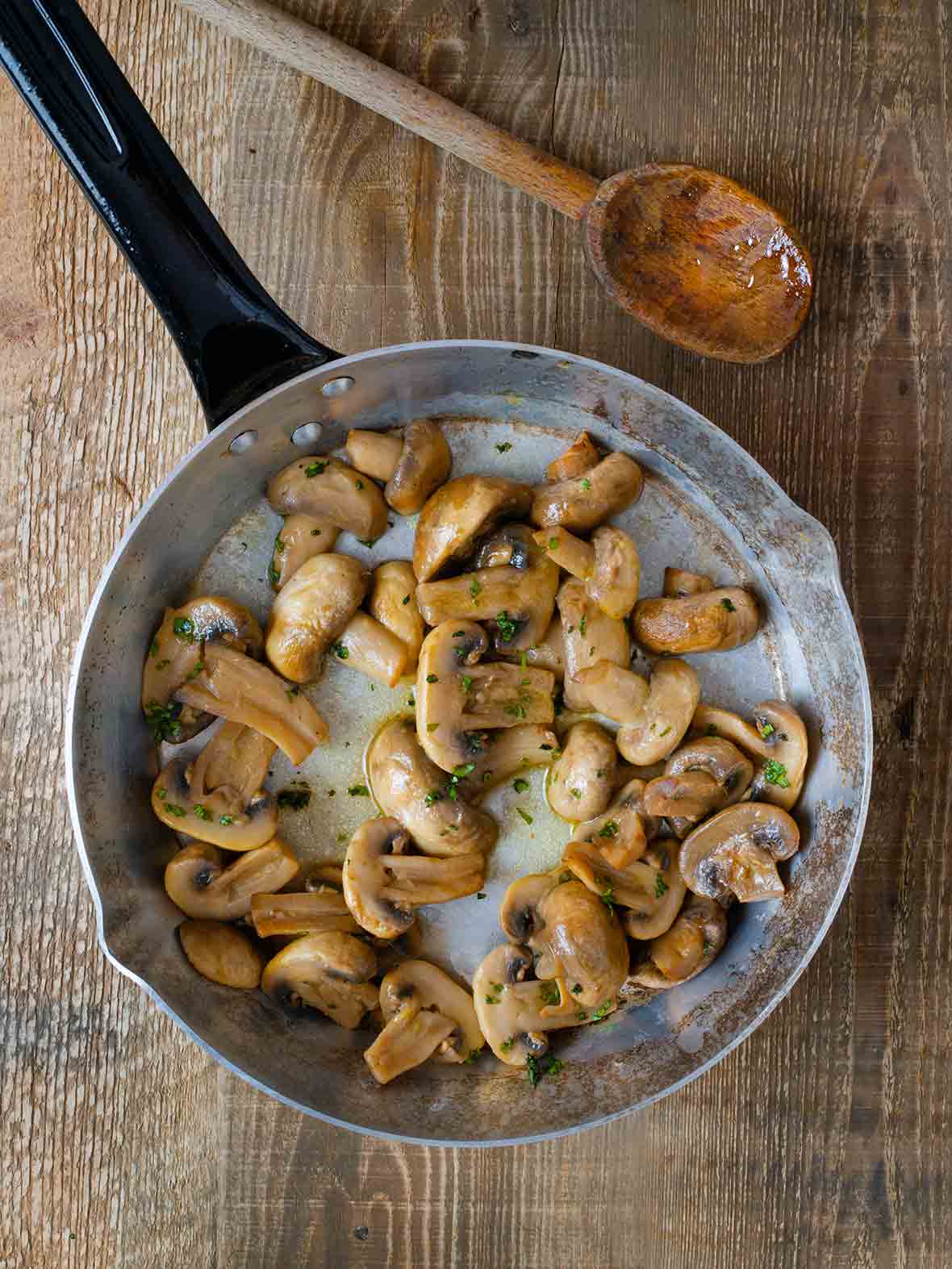 A skillet filled with mushrooms with garlic and sherry, sprinkled with parsley, and a wooden spoon resting beside it.