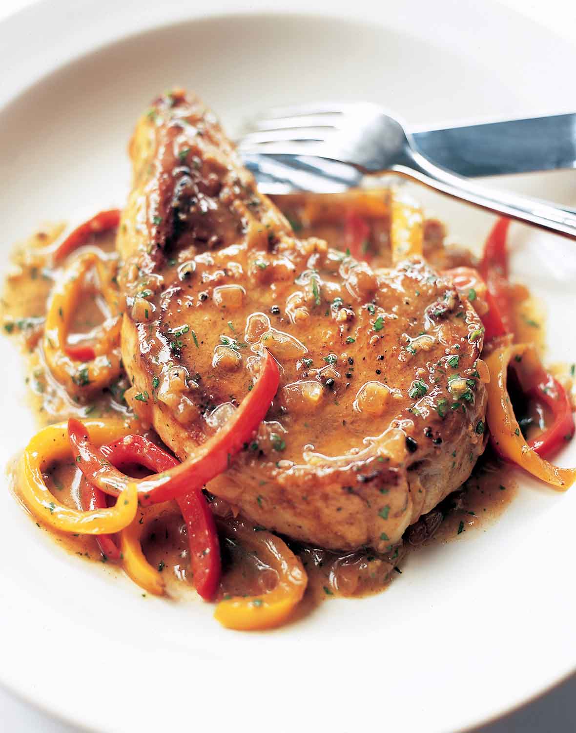 A pork chop with vinegar and sweet peppers on a white plate with a fork and knife.