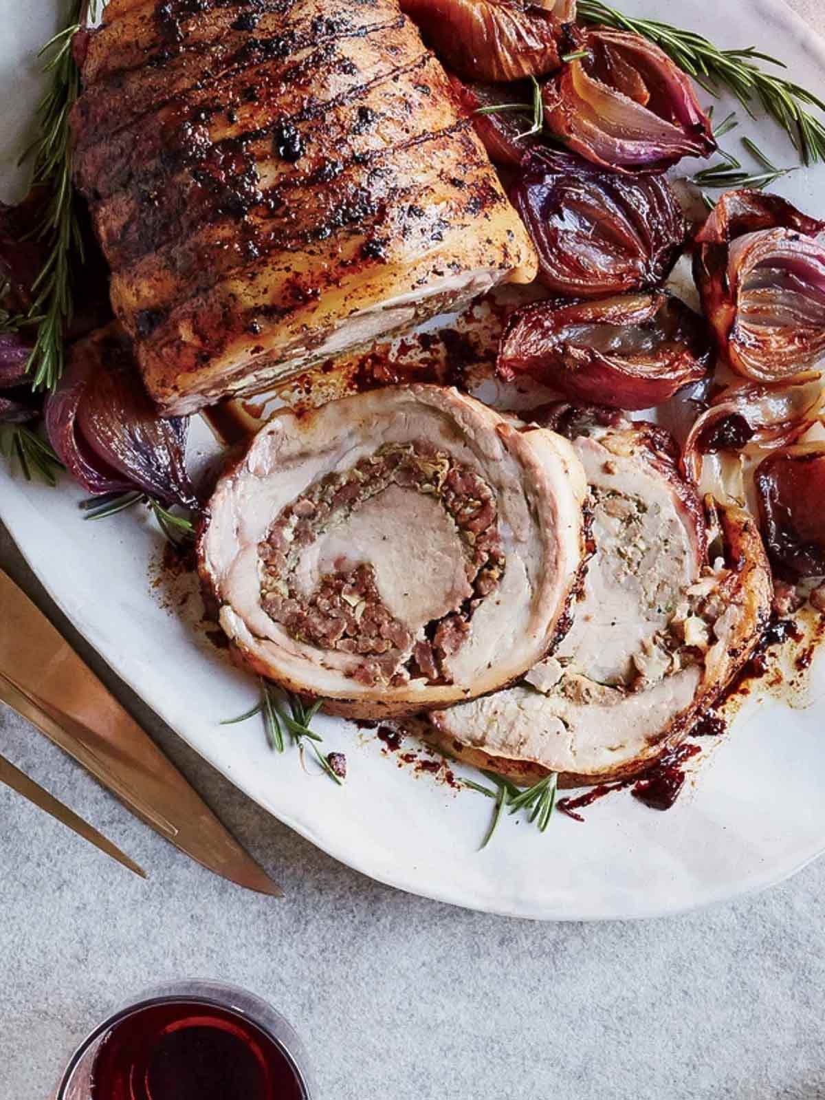 A tied pork loin in the style of Porchetta on a white platter with cooked red onions and rosemary.