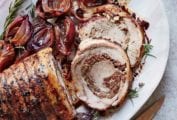 A tied pork loin in the style of Porchetta on a white platter with cooked red onions and rosemary.