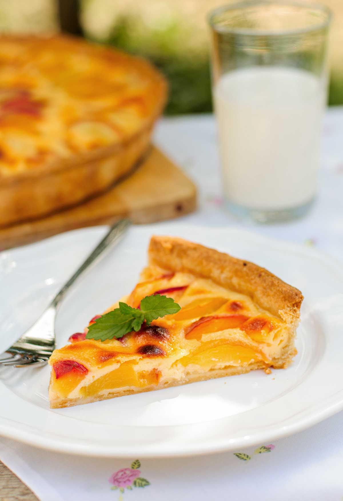 A slice of peach cream tart garnished with a mint sprig on a white plate with a fork, and a glass of milk in the background.