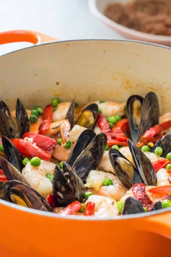 An orange Dutch oven filled with shrimp, mussels, peas, chicken, red bell peppers, on saffron rice.