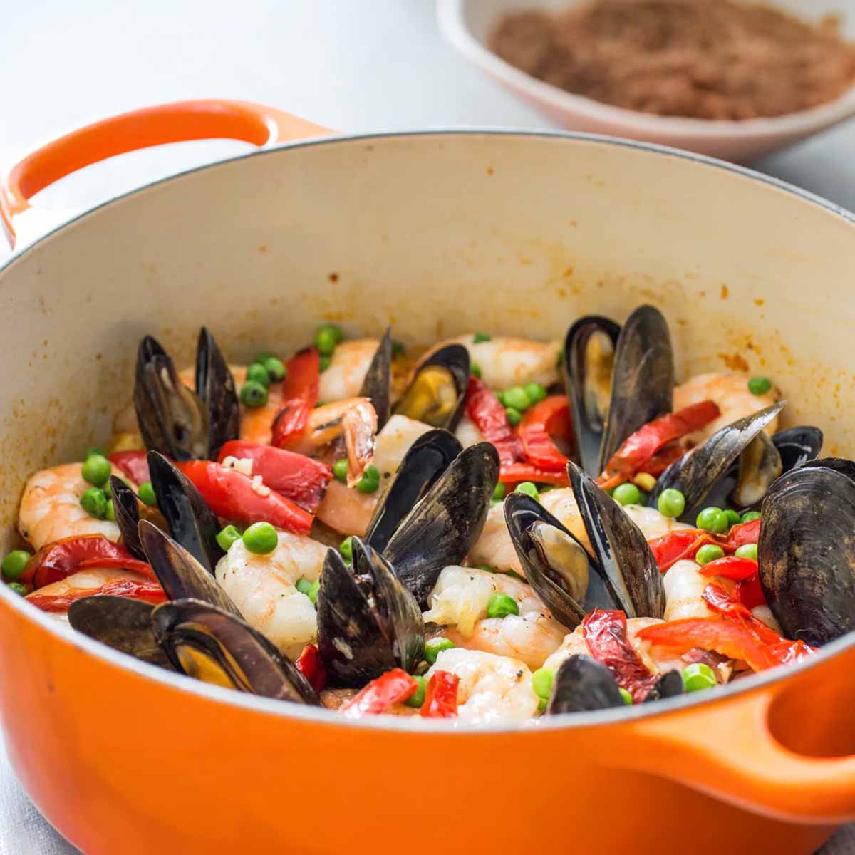 An orange Dutch oven filled with shrimp, mussels, peas, chicken, red bell peppers, on saffron rice.