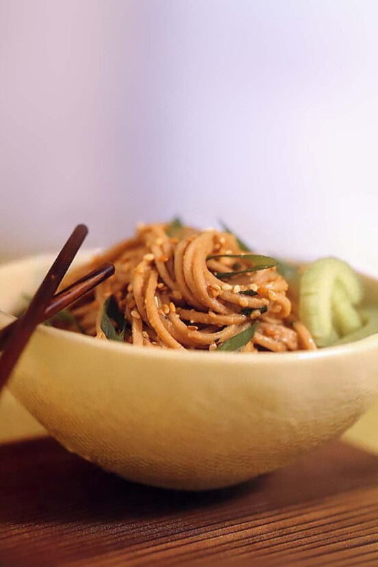 A bowl of sesame-peanut noodles coated in peanut sauce, topped with sesame seeds, chopsticks on the side.