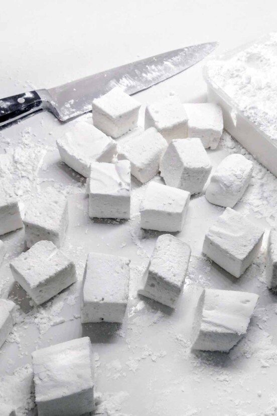 Cubes of homemade marshmallows on a white surface with a chef's knife lying beside.