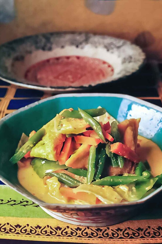 An octagonal bowl filled with tofu and vegetables in coconut milk on a colorful cloth with a large bowl in the background.