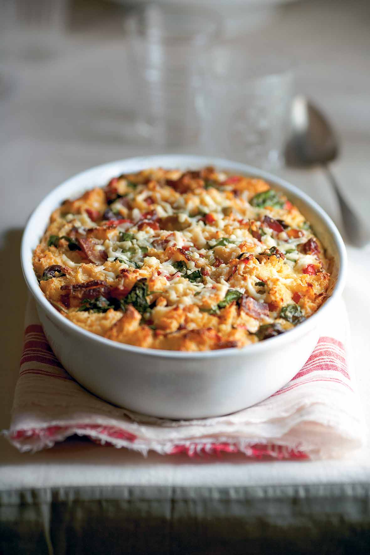 A white bowl of savory bread pudding with bacon, peppers, and spinach on top of a folded napkin