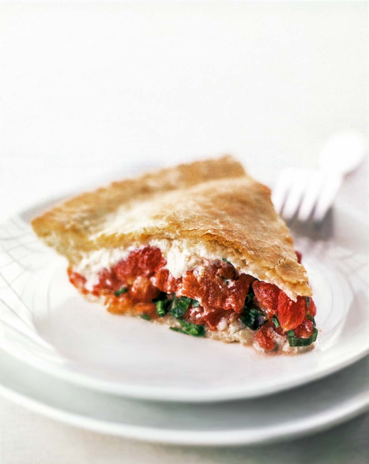 A wedge of tomato pie on a white plate with a fork in the background.
