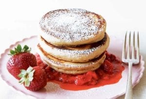 A stack of three whole-wheat pancakes with strawberry sauce, a couple whole strawberries, and confectioners' sugar sprinkled over the top.