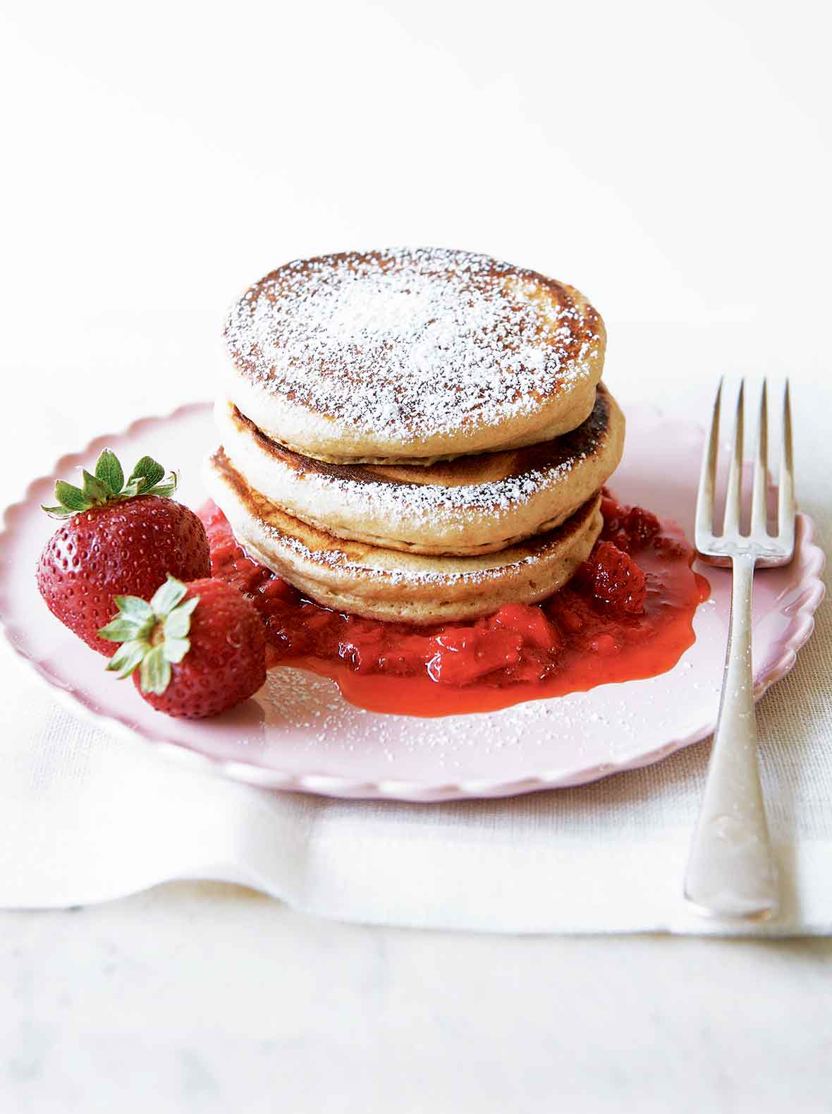 A stack of three whole-wheat pancakes with strawberry sauce, a couple whole strawberries, and confectioners' sugar sprinkled over the top.