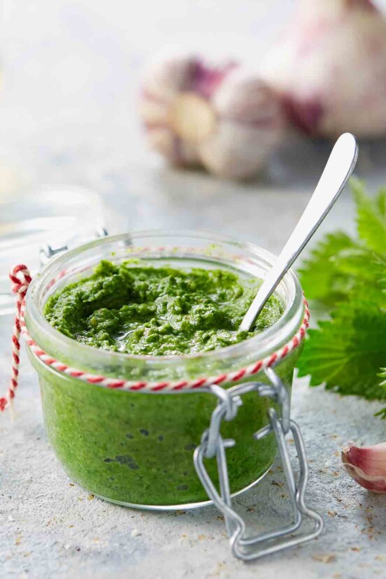 A small Mason jar of green nettle pesto with garlic cloves and nettle leaves nearby.
