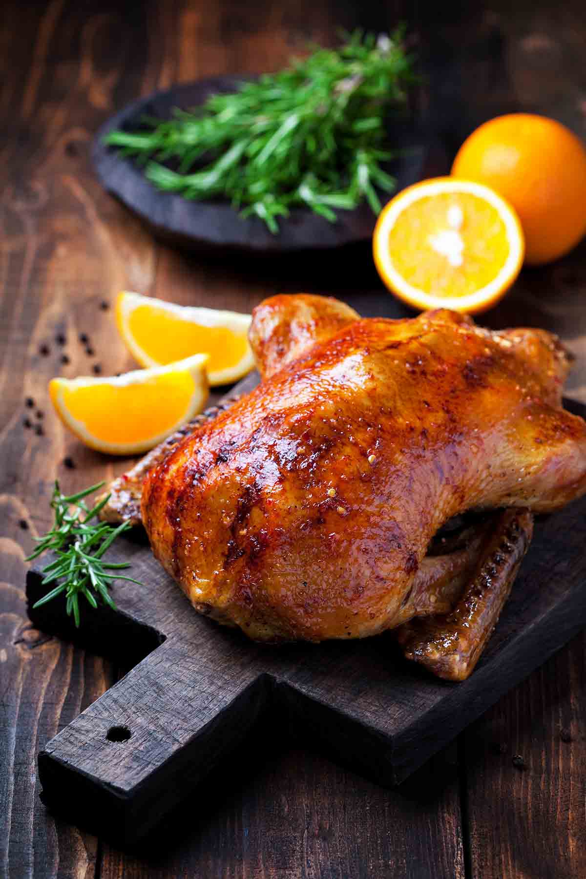 A cooked spit-roasted duck with orange and rosemary in the background.