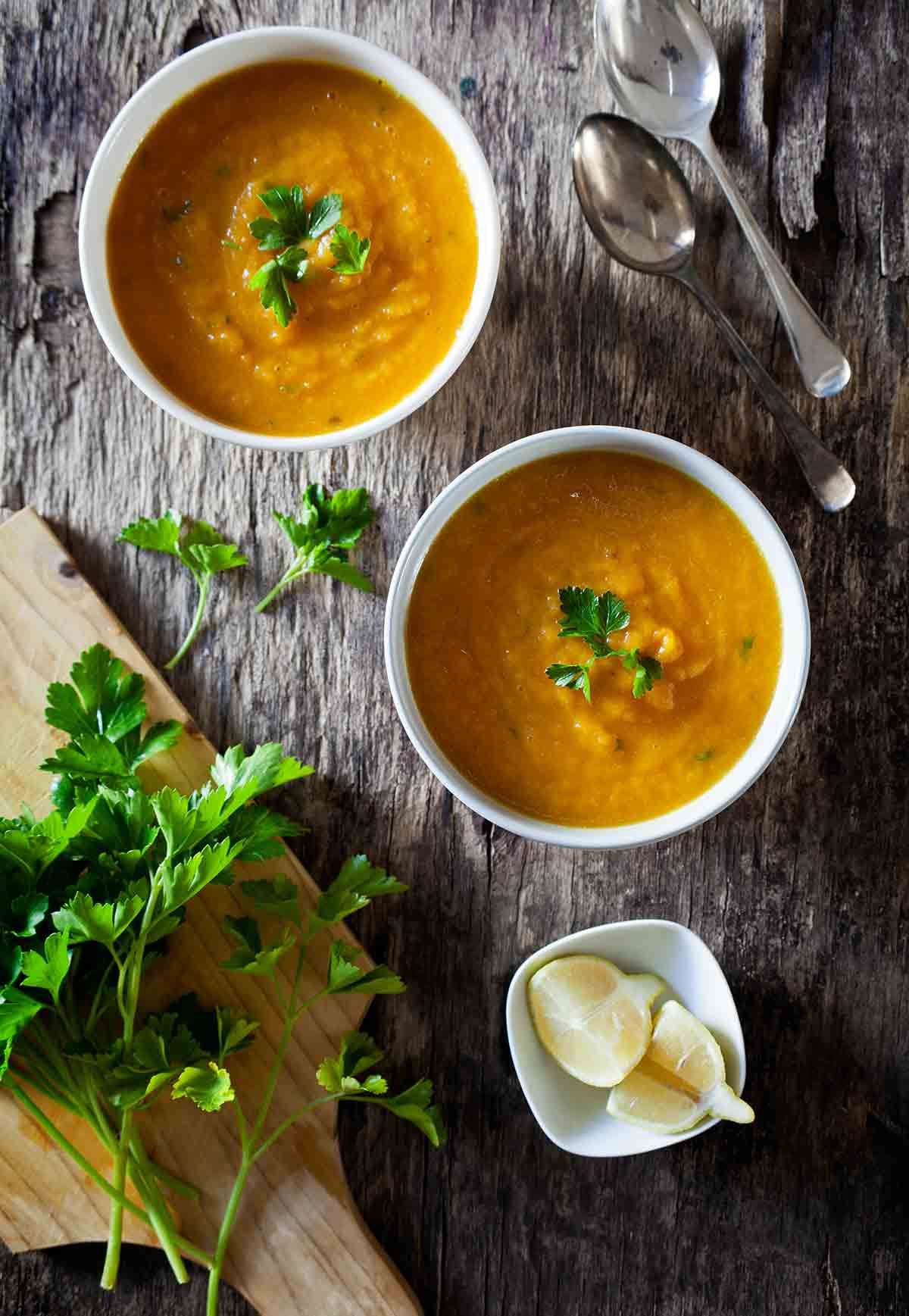 Two white bowls of carrot and ginger soup on a piece of wood