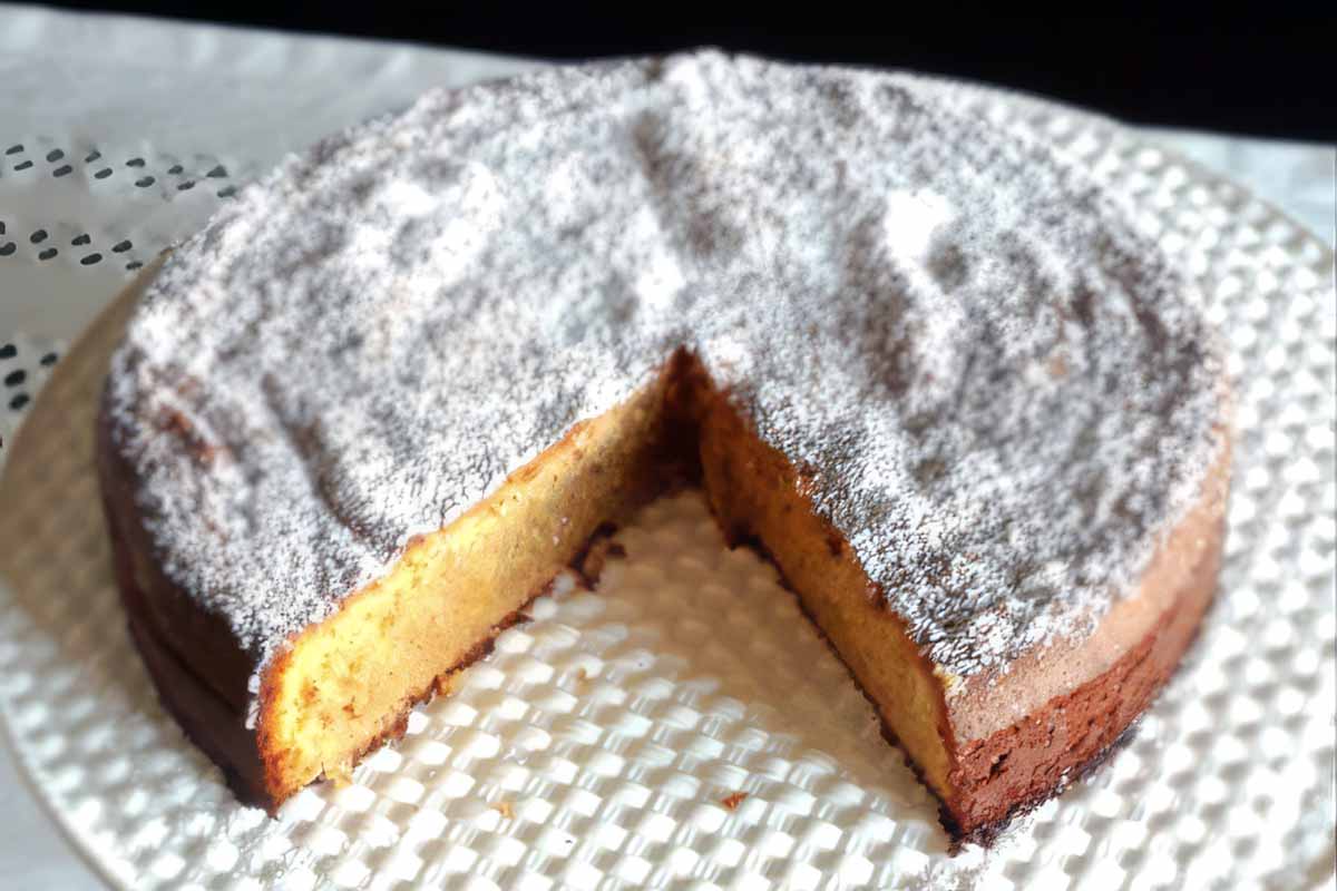 A Portuguese almond cake dusted with powdered sugar and with a big slice taken out on a white woven platter.