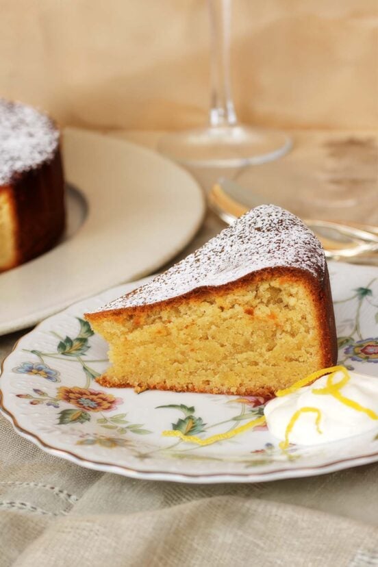 A Portuguese almond cake, often referred to as a torte, sliced on a flowered plate with dollop softly whipped cream and lemon zest.