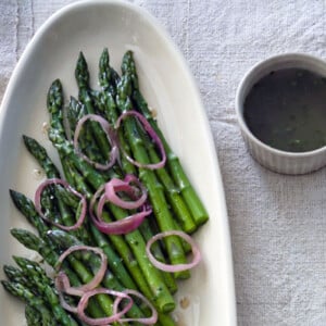 A white oval platter filled with asparagus with raspberry-shallot vinaigrette, and pickled shallots scattered on top.