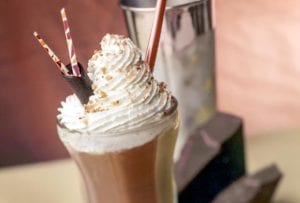 Tall glass filled with a chocolate milkshake, topped with whipped cream, chocolate shavings, three straws