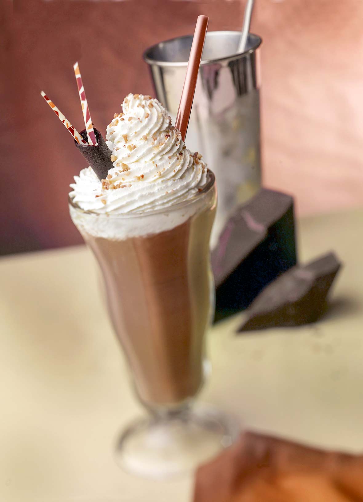 Tall glass filled with a chocolate milkshake, topped with whipped cream, chocolate shavings, three straws