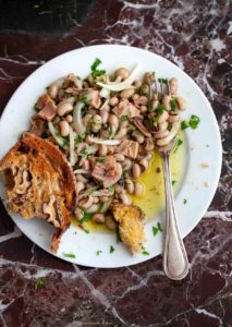 A white round plate topped with a serving of Portuguese salad of black-eyed peas with tuna with a fork resting in the salad and a crust of bread on the side.