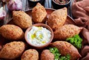A platter of Brazilian beef kibbe with a cup of yogurt mint dressing in the center and cups of pine nuts and peppercorns, and a head of garlic on the side.
