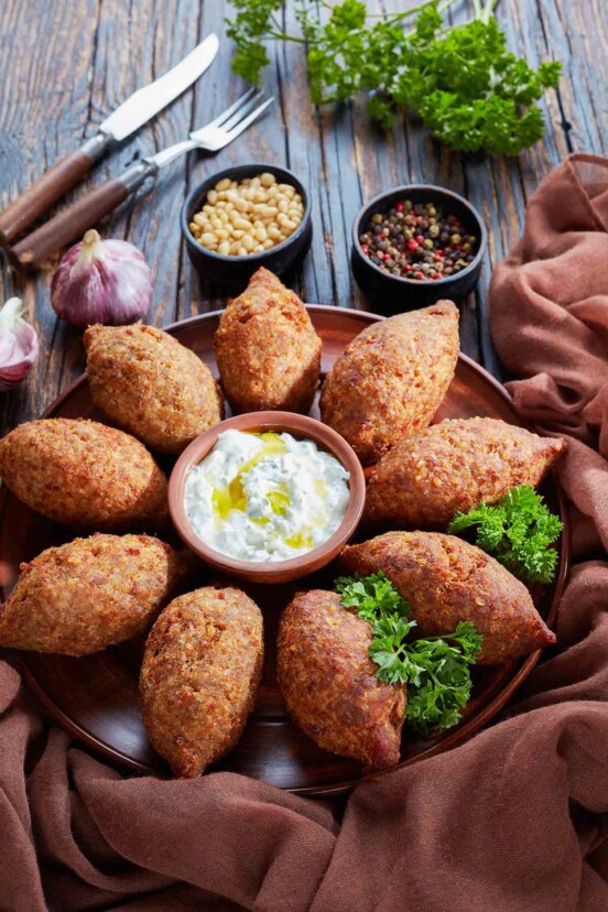A platter of Brazilian beef kibbe with a cup of yogurt mint dressing in the center and cups of pine nuts and peppercorns, and a head of garlic on the side.