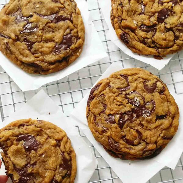 Four large chocolate chip cookies on a cooling rack with a hand reaching for one.