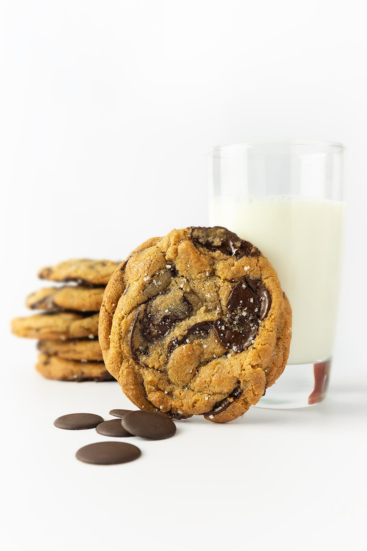A New York Times chocolate chip cookie (David Leite), leaning against a glass of milk; a stack of cookies in back.